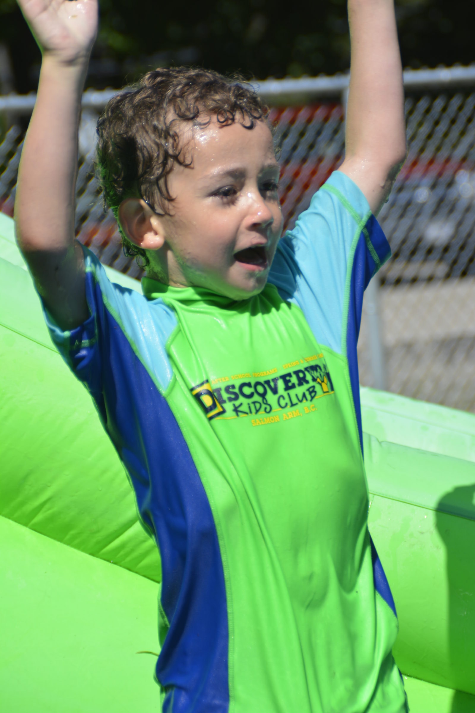 An excited Discovery Kids Club boy in a club shirt celebrating with arms raised at a water activity